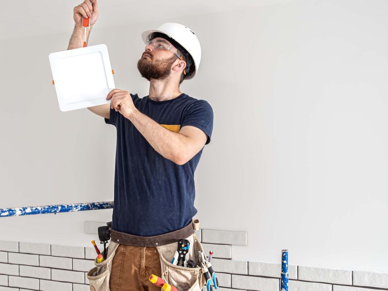 Electrician Builder with beard worker in a white helmet at work, installation of lamps at height. Professional in overalls with a drill on the background of the repair site.