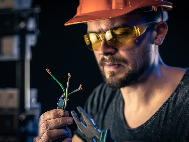 A male electrician in a protective helmet works in a switchboard with an electrical connecting cable.